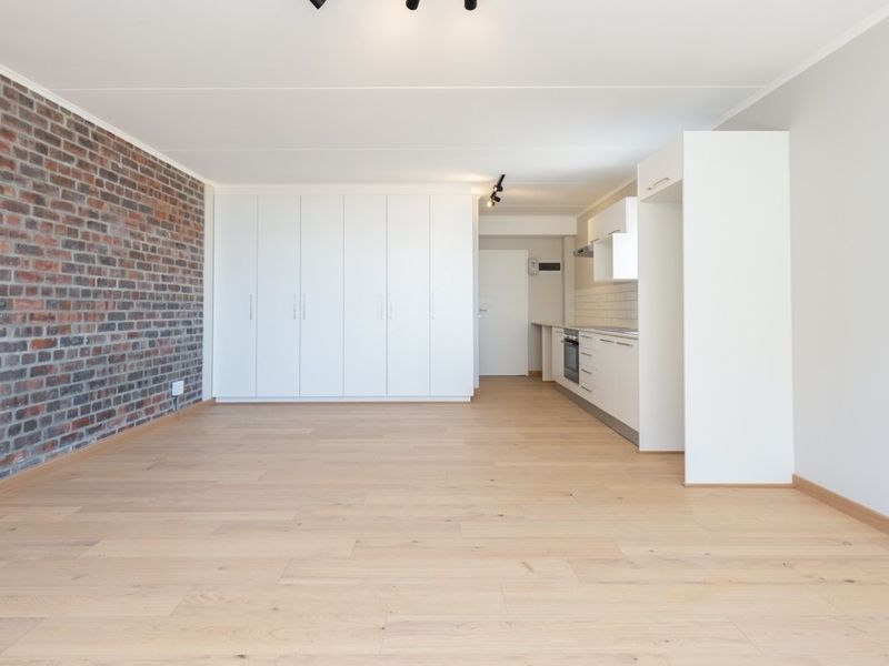 Spacious open-plan living area featuring light laminate flooring, a striking exposed brick feature wall, and a modern kitchen with built-in cabinetry and appliances.