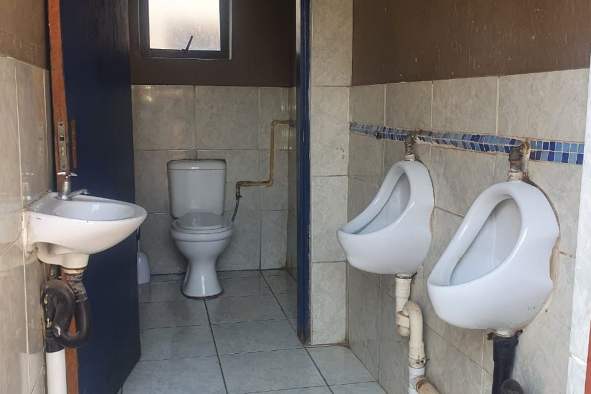 A functional bathroom featuring a flush toilet, two wall-mounted urinals, and a small washbasin with tiled walls and flooring.