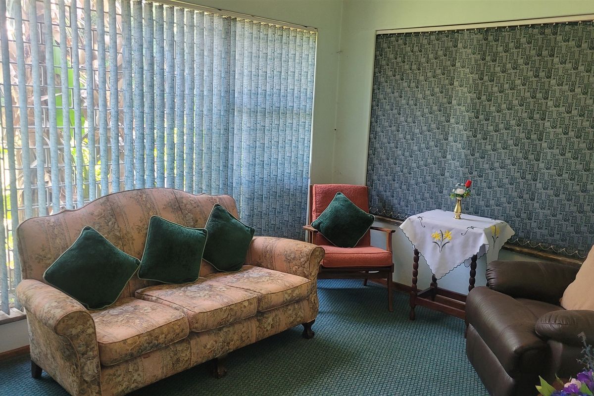 A living room area featuring fitted carpeting, vertical blinds, and patterned roller blinds, offering a dated interior space ready for modernization.