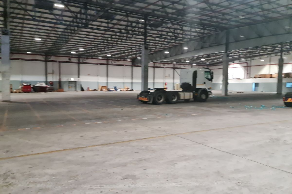 Large industrial warehouse space featuring high ceilings with exposed steel trusses, concrete flooring, and overhead industrial lighting.
