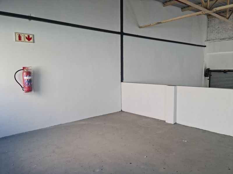 An open-plan industrial utility space featuring concrete flooring, exposed roof trusses, a roller shutter door, and wall-mounted fire safety equipment.