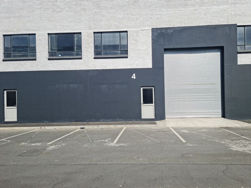Industrial unit exterior featuring a large roller shutter door, pedestrian access, and dedicated asphalt parking space in a functional commercial setting.