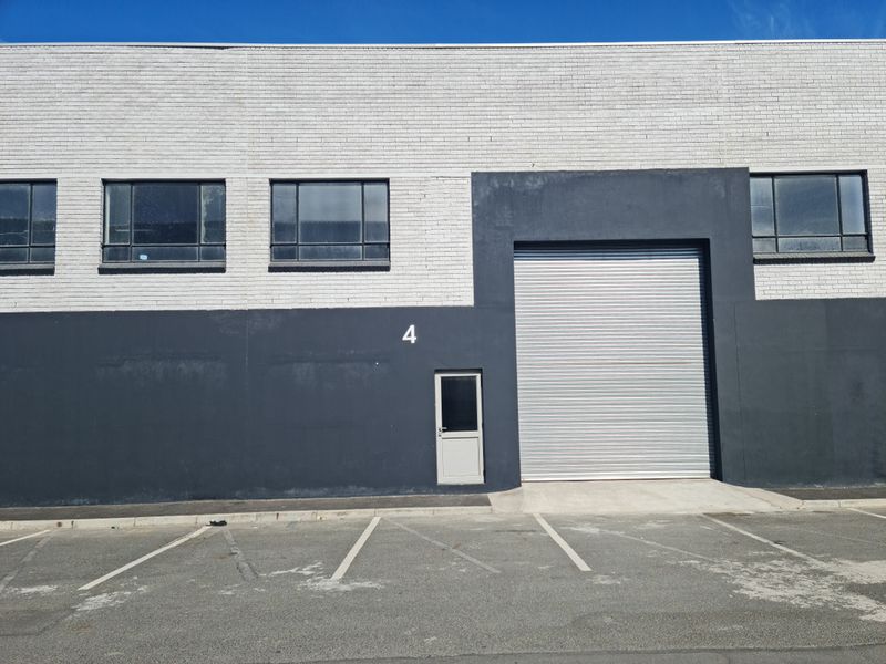 Industrial unit exterior featuring a large roller shutter door, pedestrian access door, and multiple steel-framed windows with an adjacent asphalt parking area.