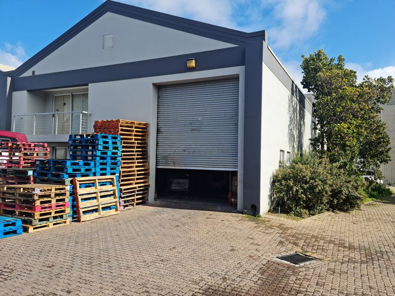 Industrial warehouse exterior featuring a large roller shutter door, paved driveway access, and a small upper-level balcony.