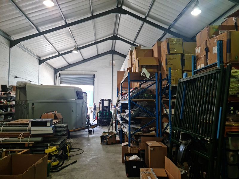 Spacious industrial warehouse unit featuring high ceilings with exposed steel trusses, concrete flooring, and a large roller shutter door for vehicle access.