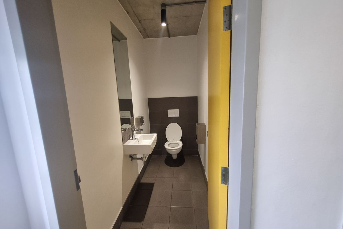 A modern industrial-style bathroom featuring a wall-mounted basin, a concealed cistern toilet, tiled flooring, and an exposed concrete ceiling with spotlighting.