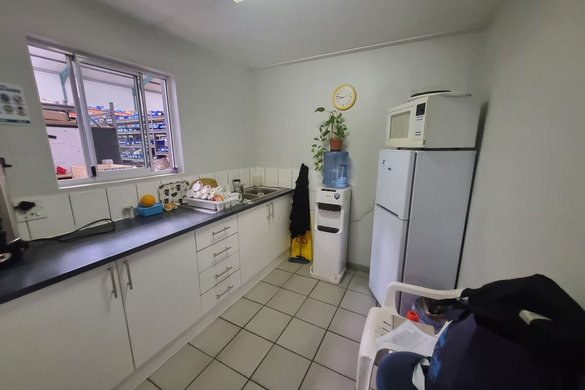 A functional industrial kitchenette featuring white cabinetry, dark countertops, a stainless steel sink, and tiled flooring, with a window overlooking the warehouse area.