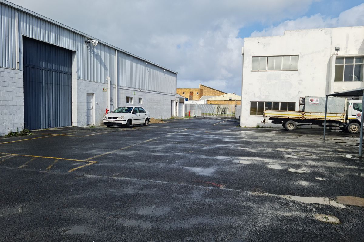 Industrial yard area featuring asphalt paving, large warehouse roller shutter door, and adjacent multi-story building facade with parking space.