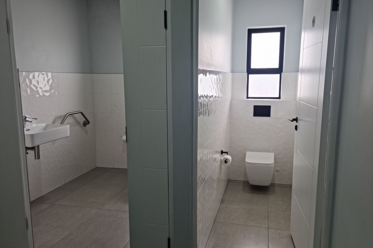 Modern industrial-style restroom facility featuring clean white textured wall tiles, grey floor tiles, wall-hung toilet, and a wall-mounted basin with grab rail.