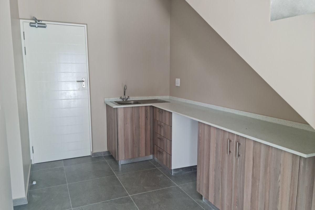 A compact kitchenette area featuring modern wood-grain cabinetry, stone-look countertops, a stainless steel sink, and tiled flooring, situated beneath a staircase.