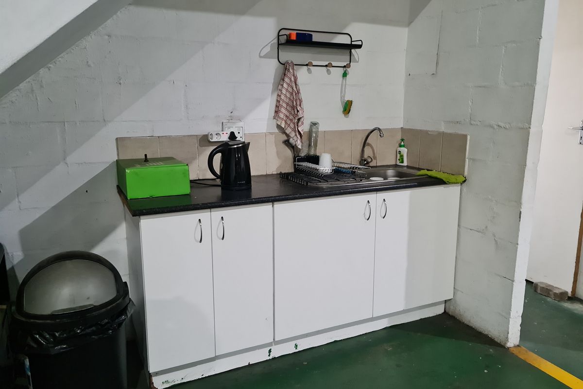 A compact kitchenette area featuring white cabinetry, a dark countertop, a stainless steel sink, and tiled splashback, situated under a staircase.
