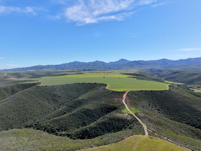 An aerial view of the expansive farm property featuring rolling hills, cultivated green fields, and a dirt access road against a mountain backdrop.