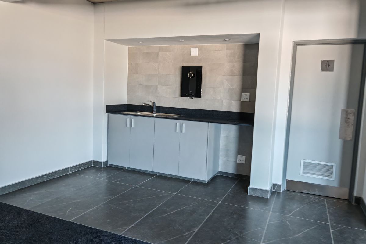 A clean kitchenette area featuring grey cabinetry, a dark countertop, a stainless steel sink, and tiled splashback, adjacent to a restroom door.