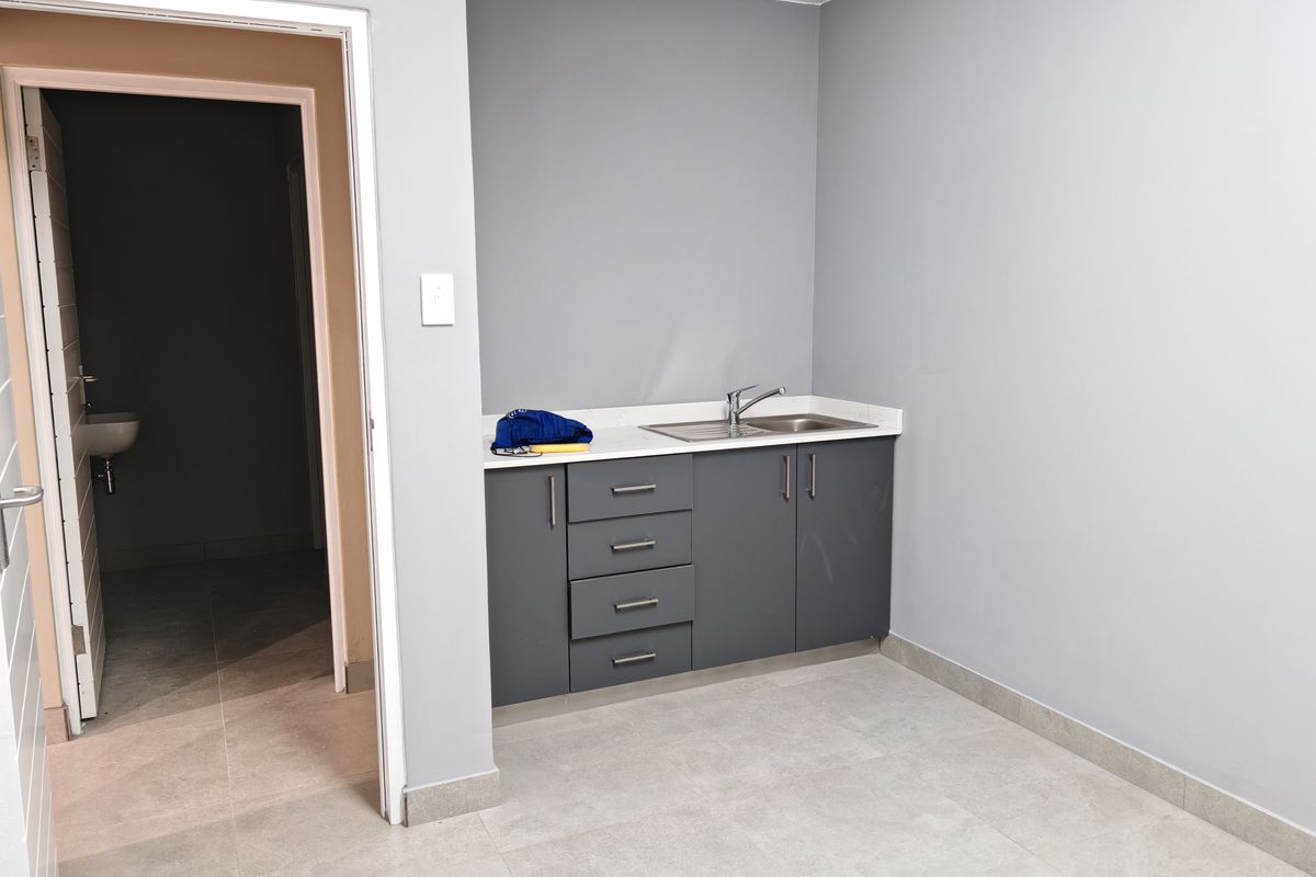 A functional kitchenette area featuring dark grey cabinetry, a stainless steel sink, and light-toned tiled flooring, with an adjacent doorway leading to a restroom.