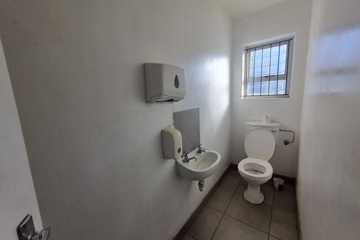 A functional industrial restroom featuring a wall-mounted basin, toilet, and security-barred window, finished with tiled flooring and basic wall-mounted dispensers.
