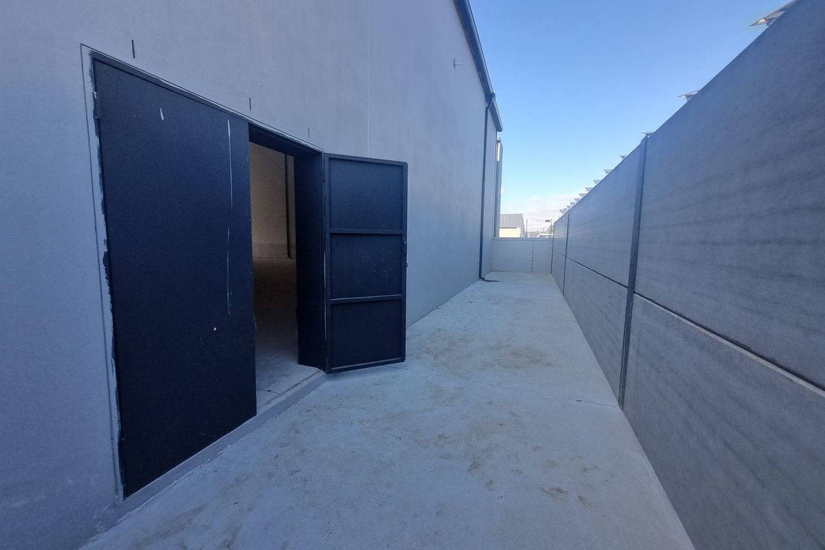 A side exterior view of an industrial property featuring a concrete walkway, a secure metal door, and a perimeter concrete wall.