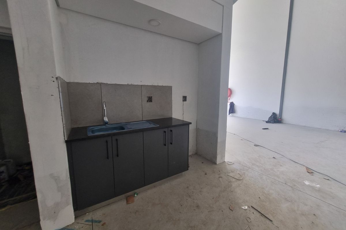 A compact kitchenette area featuring dark cabinetry, a stainless steel sink, and tiled splashback, situated within an open-plan industrial warehouse space.