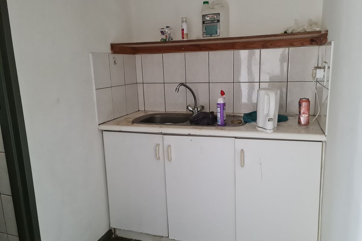 A compact kitchenette area featuring a stainless steel sink, white base cabinetry, and tiled splashback with an overhead wooden shelf.