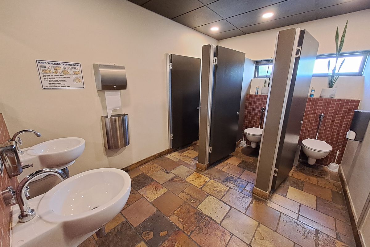 A functional office restroom featuring dual wall-mounted basins, multiple toilet cubicles with privacy partitions, and durable stone-look floor tiling.