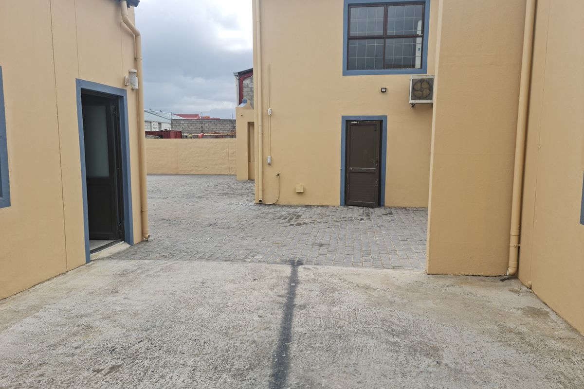 A paved industrial courtyard area situated between building structures, featuring concrete flooring, exterior access doors, and wall-mounted air conditioning unit.