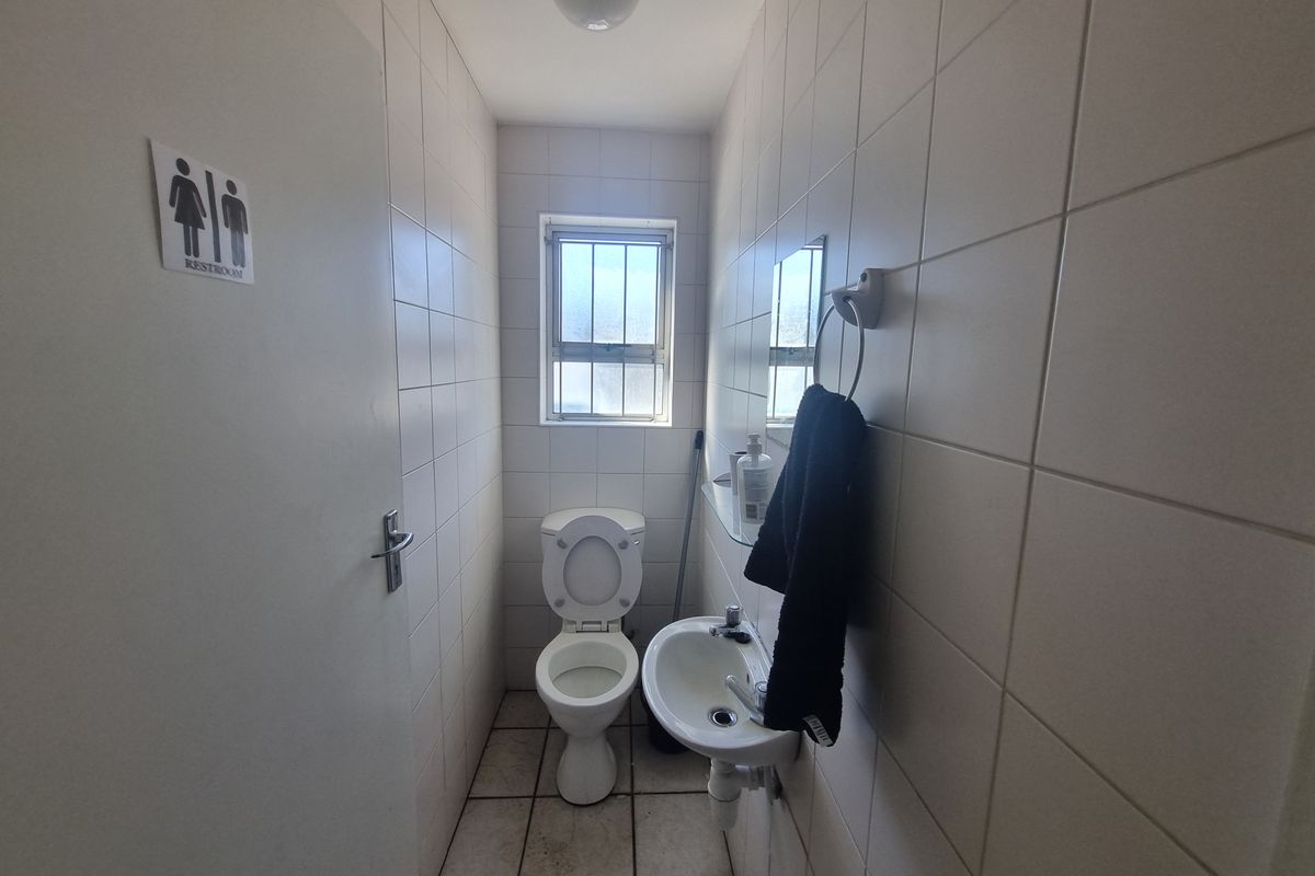 A compact industrial restroom featuring full-height wall tiling, a wall-mounted basin, and a standard toilet suite with a security-barred window.