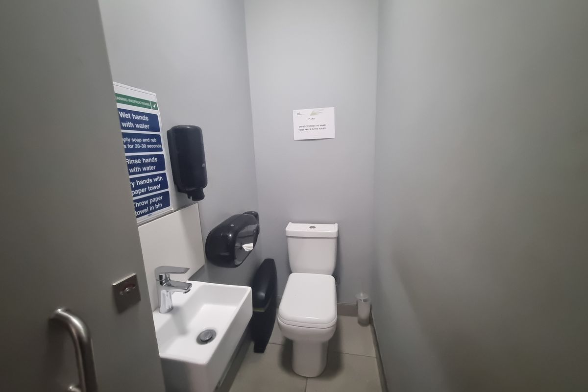 A compact industrial restroom featuring a wall-mounted basin, modern toilet, and essential hygiene dispensers.