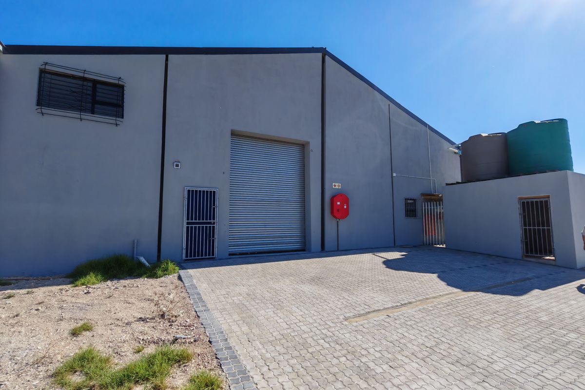 Exterior view of an industrial warehouse featuring a large roller shutter door, security gates, paved driveway, and external water storage tanks.