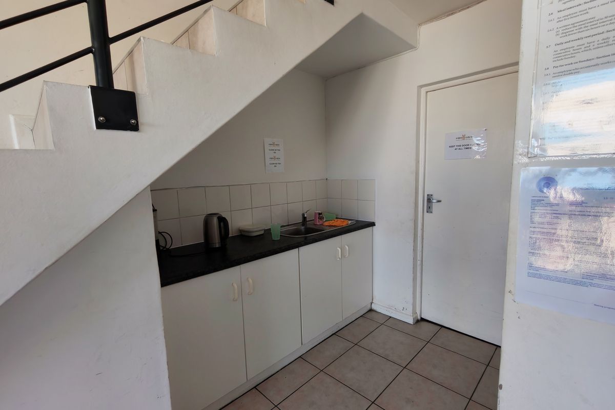A compact kitchenette area located beneath a staircase, featuring built-in cabinetry, a sink, and tiled flooring in an industrial setting.