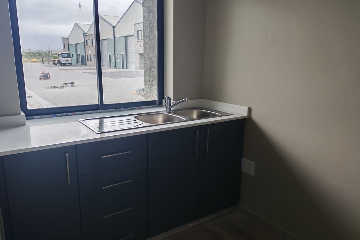 A functional kitchenette area featuring dark cabinetry, a stainless steel double sink with mixer tap, and a white countertop, overlooking an industrial complex.