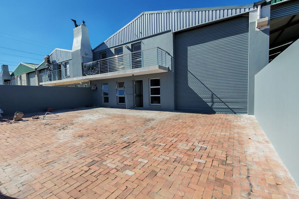 Industrial property exterior featuring a large roller shutter door, brick paved yard, and an upper-level balcony with metal railings.