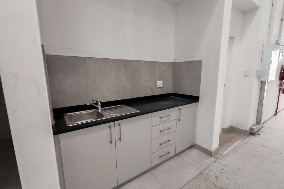 A functional kitchenette area featuring light grey cabinetry, a black countertop, stainless steel sink, and grey tiled splashback within an industrial space.