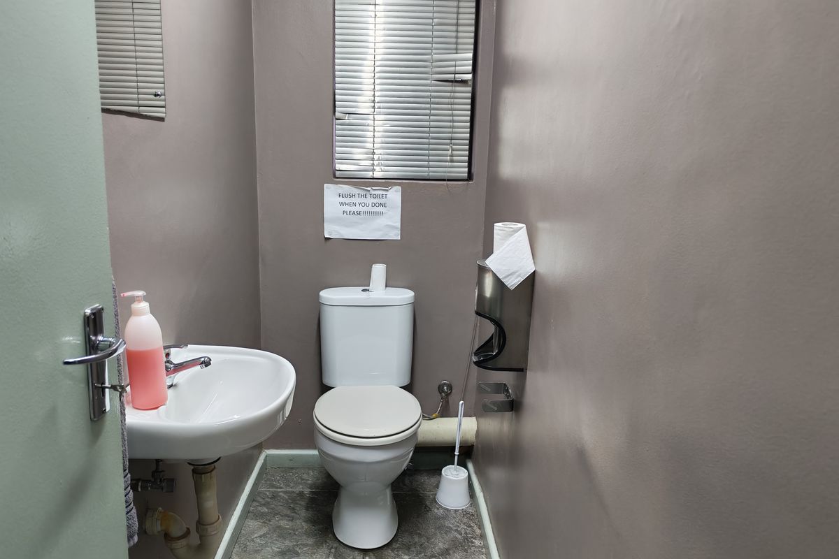 A functional industrial restroom featuring a wall-mounted basin, a standard toilet, and a window with fitted blinds.