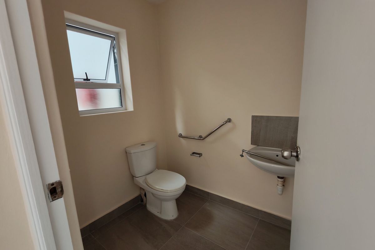 A functional, accessible restroom featuring a toilet, wall-mounted basin, and safety grab rails, finished with neutral wall paint and tiled flooring.