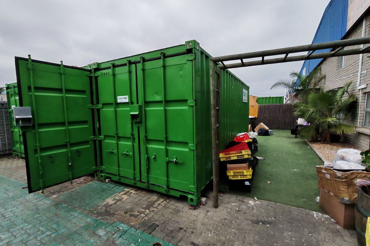 Exterior view of a green industrial storage container situated on a paved area with adjacent artificial turf and structural framing.
