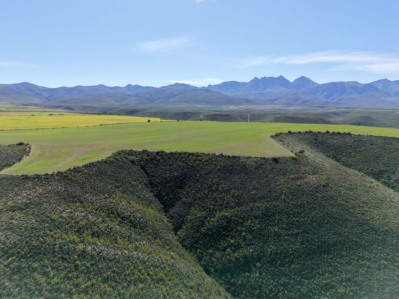 An expansive aerial view of the farm landscape, featuring rolling green fields, natural vegetation, and distant mountain ranges under a clear blue sky.