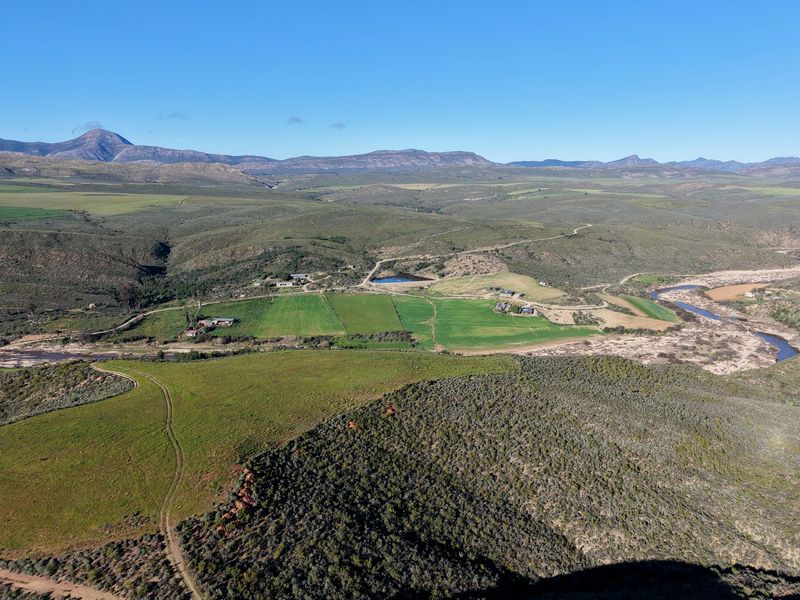 An expansive aerial view of the farm property, showcasing vast agricultural fields, natural river frontage, a dam, and surrounding mountainous landscape.