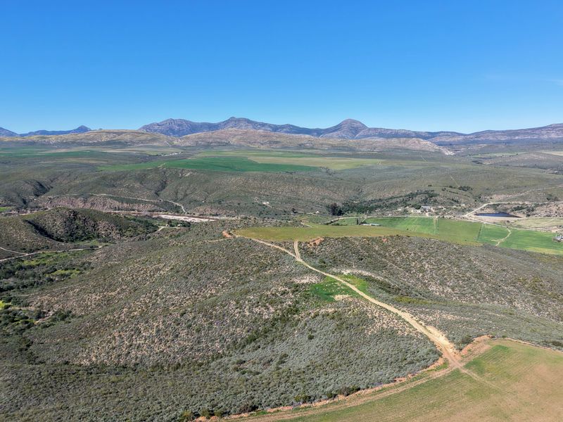 An expansive aerial view of the farm property, showcasing rolling hills, dirt access roads, and distant mountain ranges under a clear blue sky.