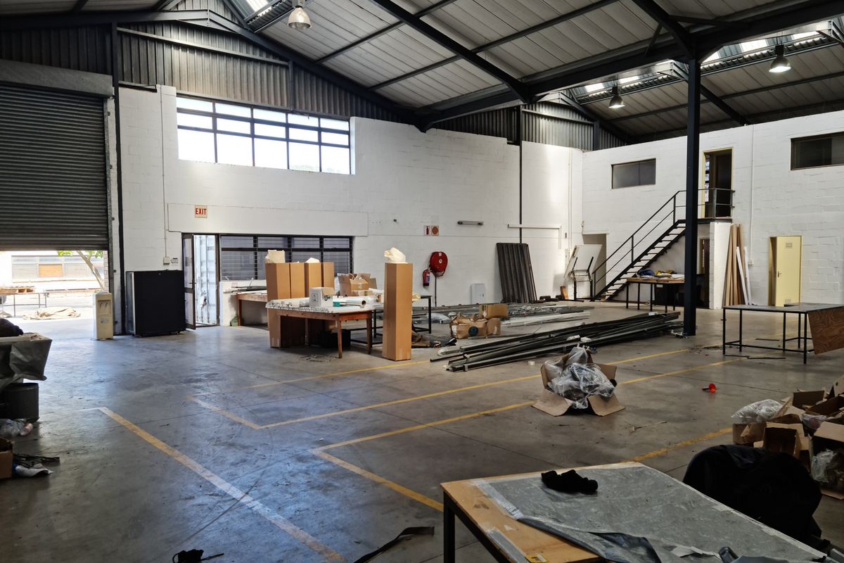 Spacious industrial warehouse unit featuring high ceilings with exposed steel trusses, skylights, concrete flooring, and a metal staircase leading to an upper level.