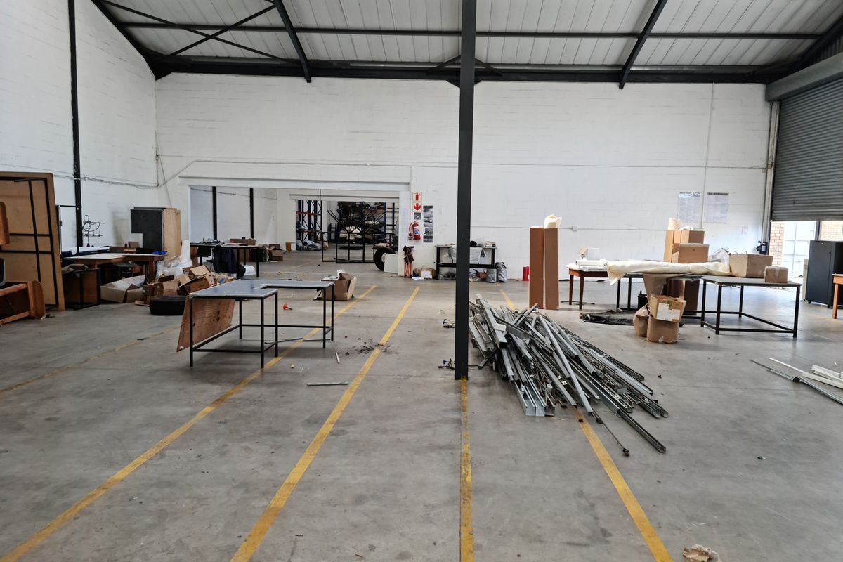 Spacious industrial warehouse unit featuring high ceilings with exposed steel trusses, concrete flooring, and a large roller shutter door for vehicle access.