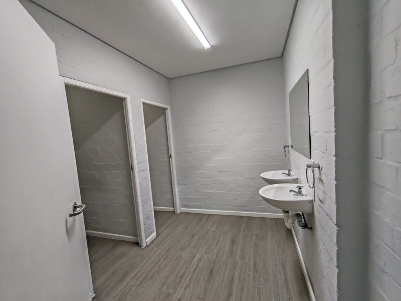 A clean, minimalist office bathroom featuring dual wall-mounted washbasins, modern laminate flooring, and painted brick walls.