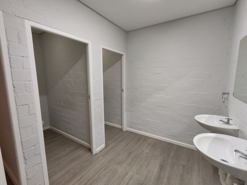 A clean, modern office bathroom featuring dual wall-mounted washbasins, painted brick walls, and durable laminate flooring with two separate cubicle entrances.