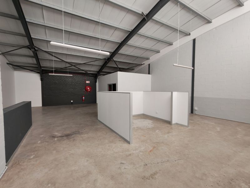 Open-plan office space featuring exposed steel roof trusses, concrete flooring, and modular partition walls, suitable for a flexible workspace layout.
