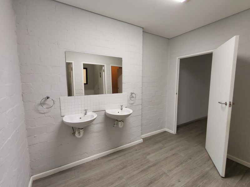 A clean, modern office bathroom featuring dual wall-mounted washbasins, a tiled splashback, and durable laminate flooring.