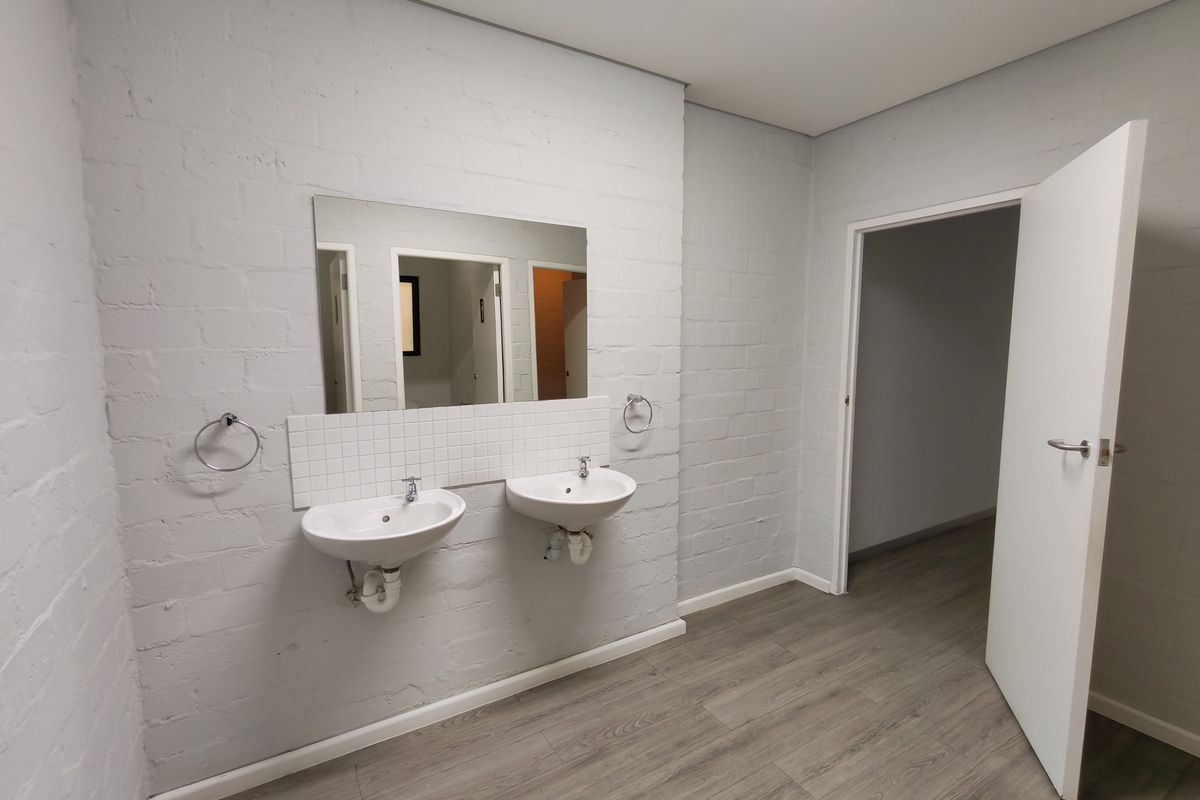 A clean, modern office bathroom featuring dual wall-mounted washbasins, a tiled splashback, and durable laminate flooring.