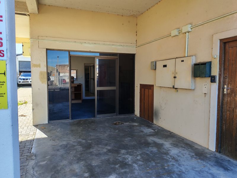 Covered industrial entrance area featuring a sliding glass door, external electrical distribution board, and concrete flooring with visible surface wear.