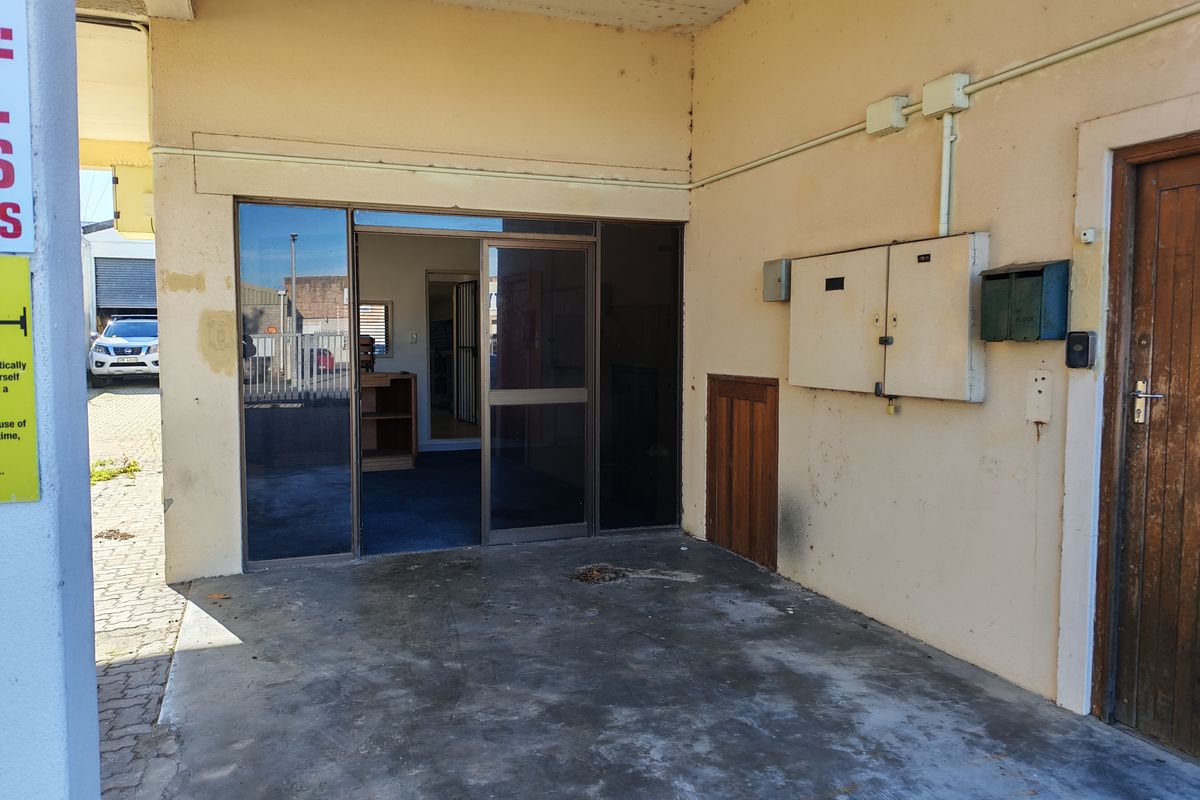 Covered industrial entrance area featuring a sliding glass door, external electrical distribution board, and concrete flooring with visible surface wear.