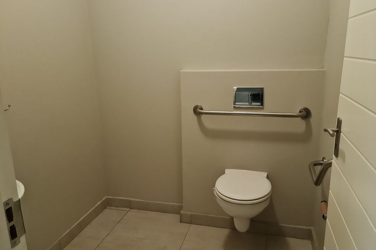 A clean, accessible bathroom featuring a wall-mounted toilet, chrome flush plate, and stainless steel grab rail with neutral tiled flooring.