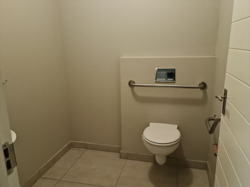 A clean, accessible bathroom featuring a wall-mounted toilet, chrome flush plate, and stainless steel grab rail with neutral tiled flooring.