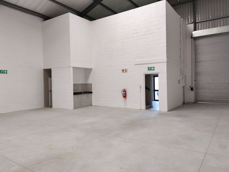 Spacious industrial warehouse unit featuring high ceilings with exposed steel trusses, concrete flooring, a roller shutter door, and a small built-in kitchenette area.