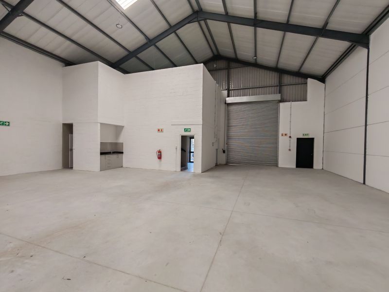 Spacious industrial warehouse unit featuring high ceilings with exposed steel trusses, concrete flooring, a roller shutter door, and a small integrated kitchenette area.